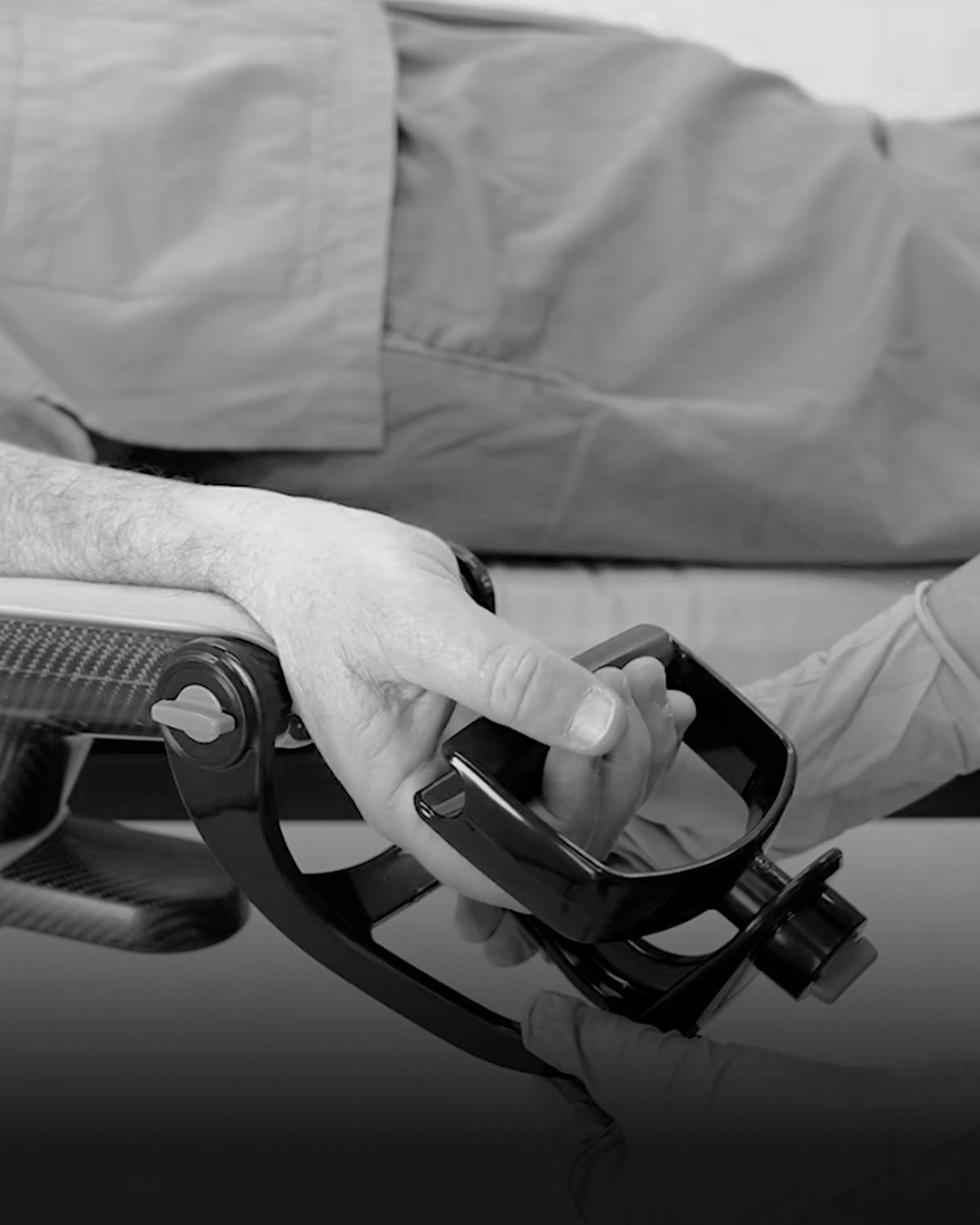 A patient's hand gripping a black medical device, with a gloved hand also on the device, in black and white.