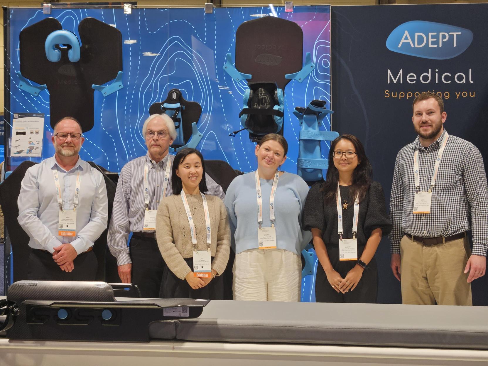 Six smiling people stand in front of an ADEPT Medical booth displaying blue medical devices.