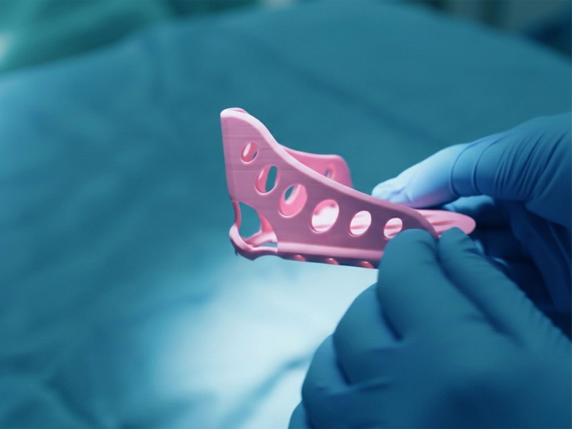 A person in blue surgical gloves holds a pink 3D-printed medical device.